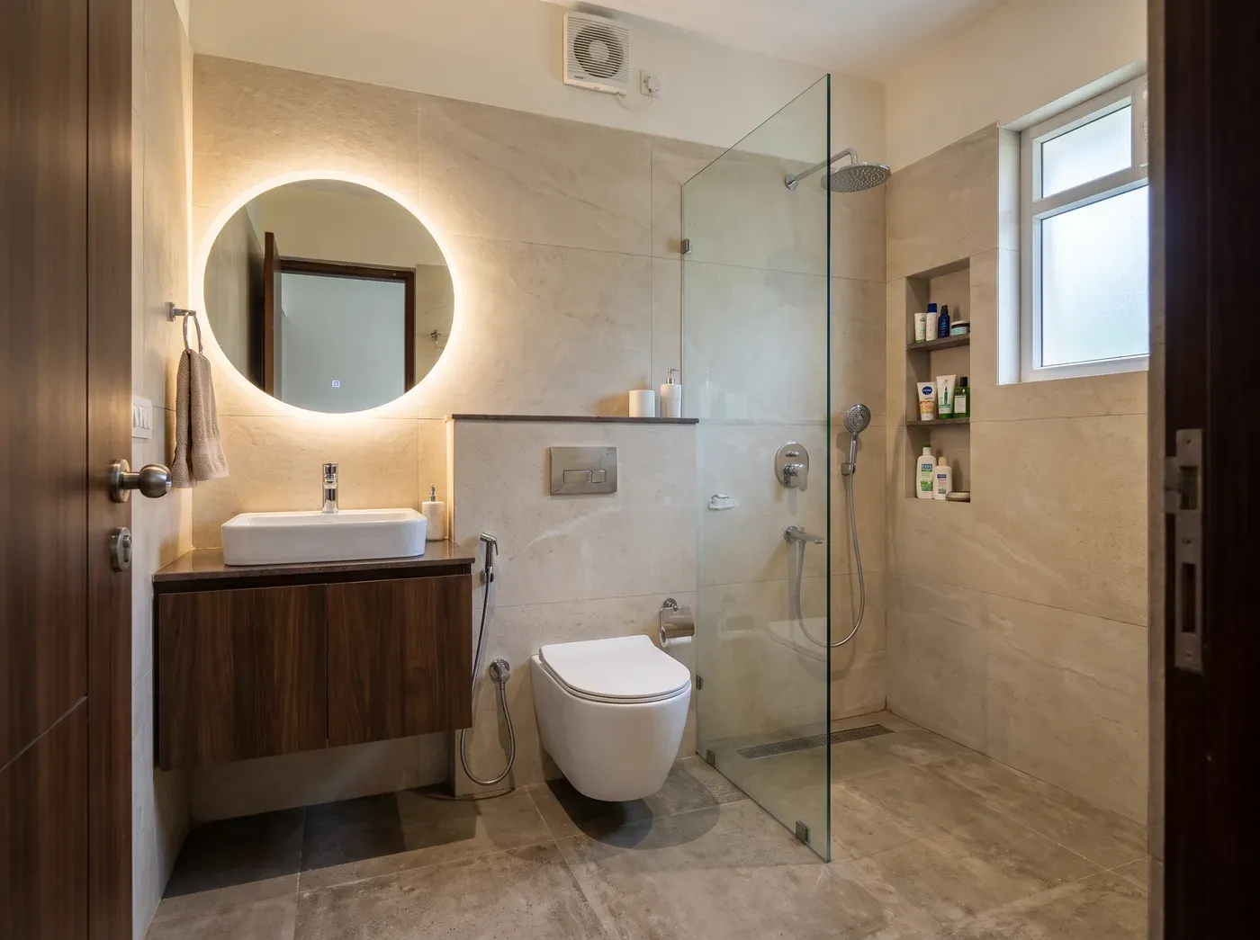 The same bathroom after renovation: large stone-look tiles, a floating vanity with lit mirror, a glass-separated shower and organised storage.