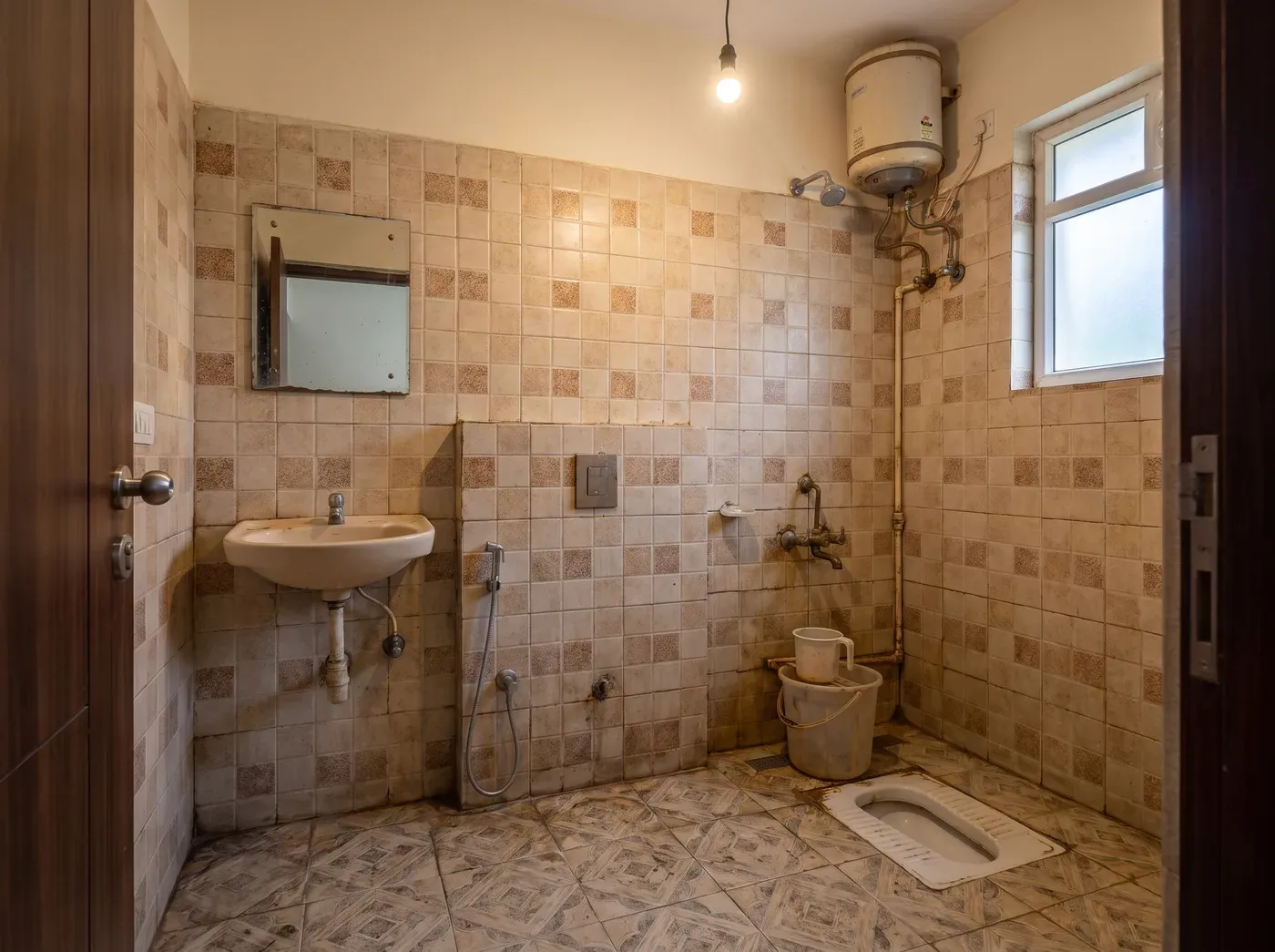 The same bathroom before renovation: a traditional Indian squat toilet, dated patterned wall tiles, a wall-hung basin, an old geyser with exposed pipework and no shower separation.