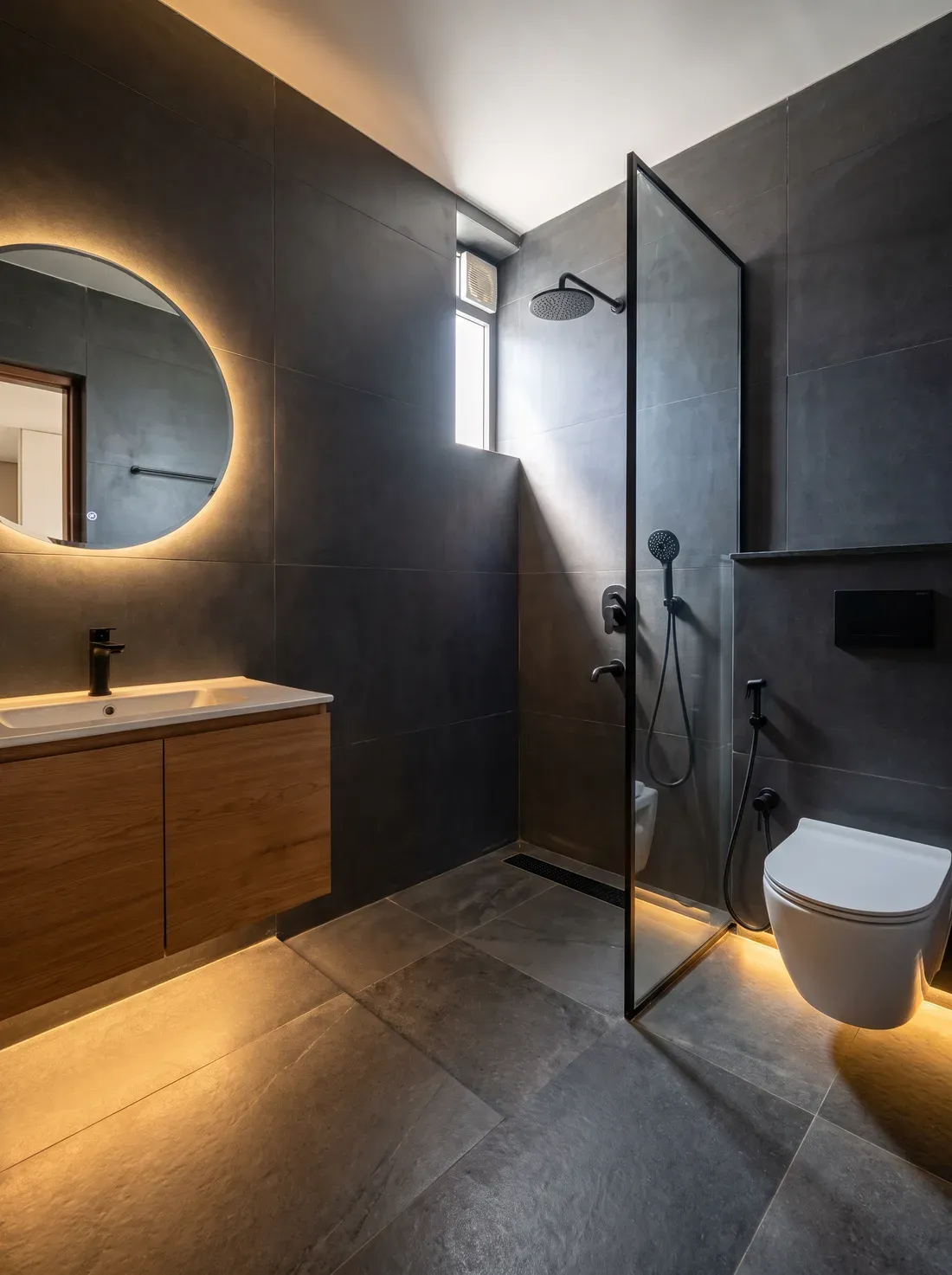 A compact apartment bathroom finished in matte charcoal tiles with a warm oak vanity, backlit round mirror, black fittings and a glass walk-in shower — a design visualisation.