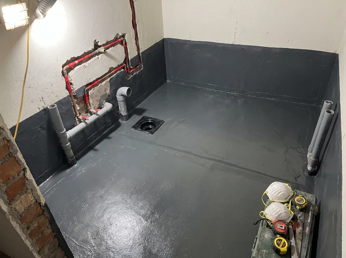 A bathroom mid-renovation before tiling: a grey waterproofing membrane coats the floor and lower walls, sealed around the floor drain and around roughed-in copper water lines and PVC drain pipes — an illustration of the hidden work behind a finished bathroom.
